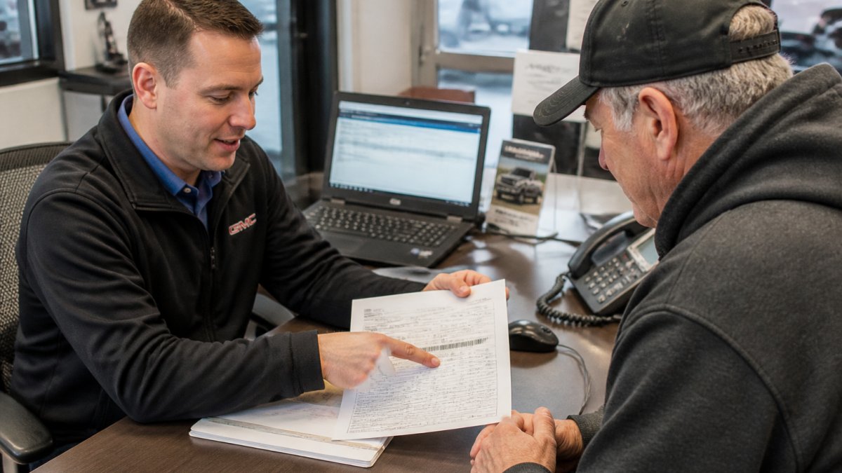A service advisor at a dealership desk pointing to a VIN printed on a vehicle registration document while a truck owner looks on, with a laptop open showing a warranty lookup screen in the background