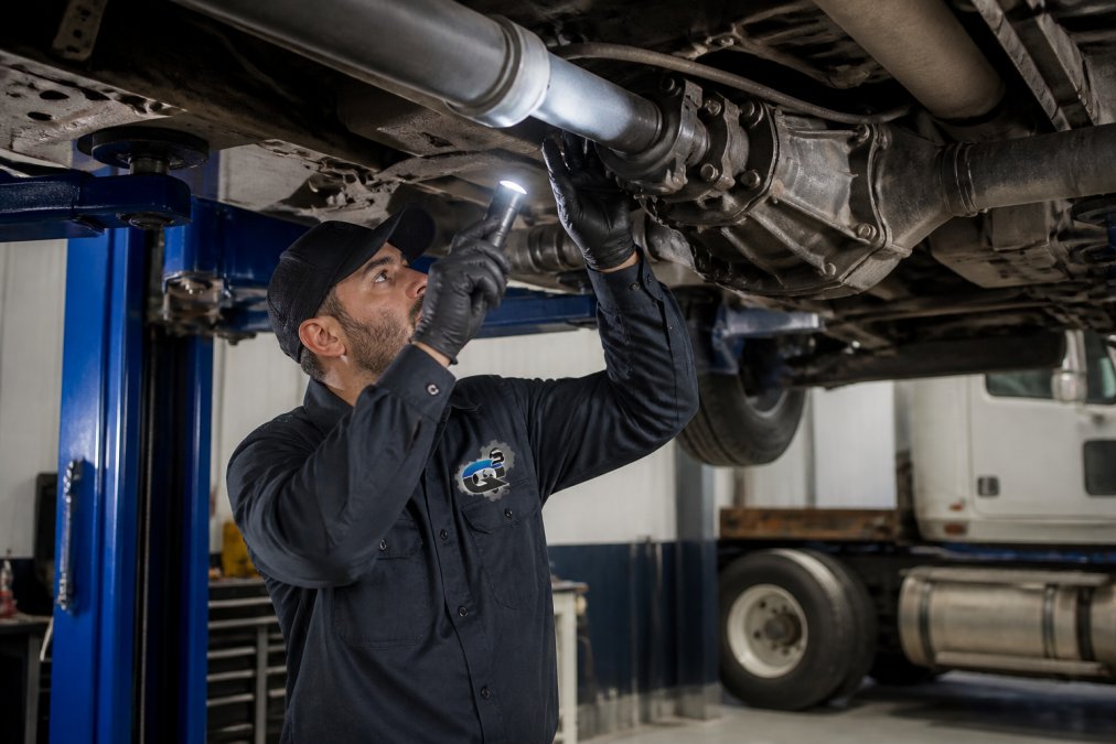 A technician in a clean dark uniform inspecting the underside of a diesel truck raised on a service bay lift, holding a flashlight and checking the driveshaft and differential