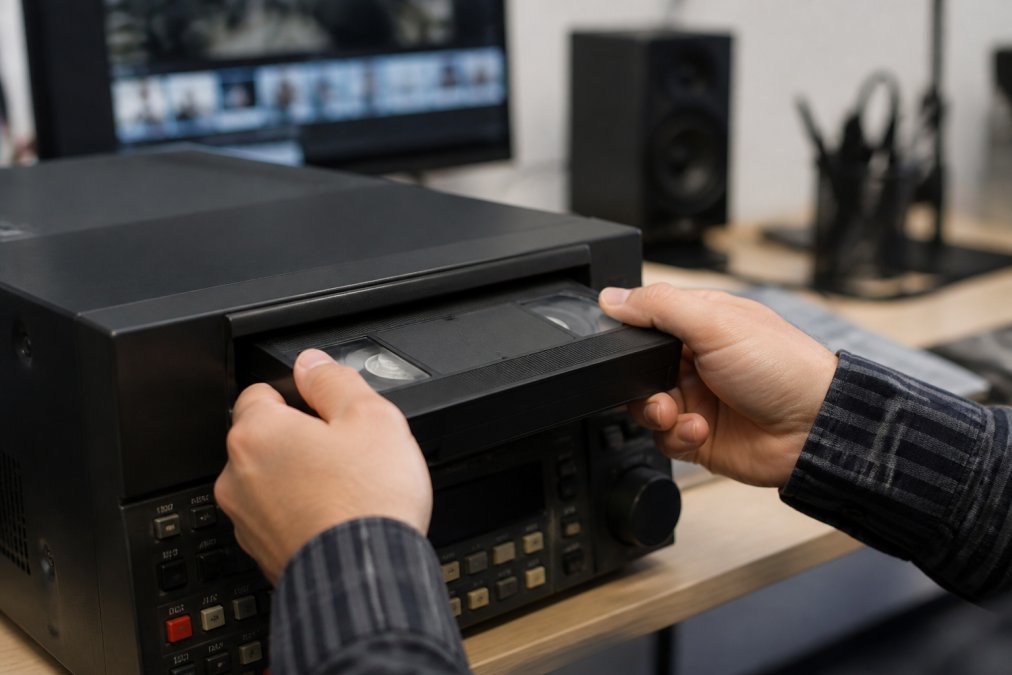 Close-up of a technician's hands carefully threading a VHS tape into a professional-grade video capture deck in a clean studio workspace, with a computer monitor showing a video timeline in the background