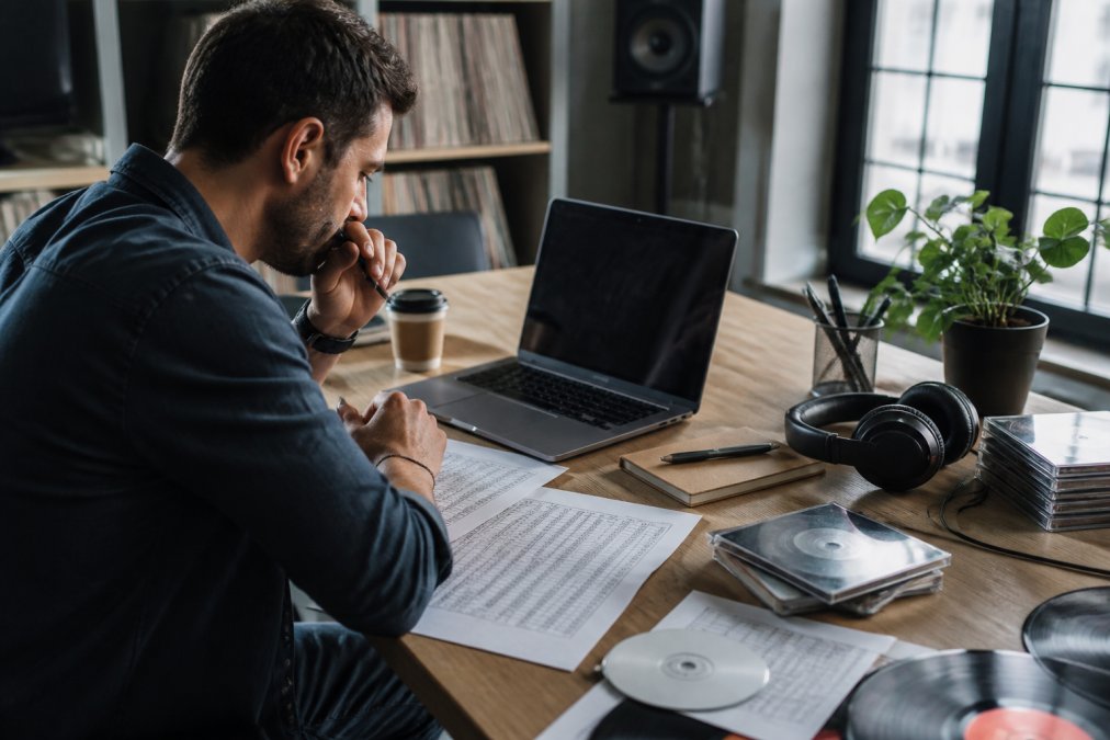 Label manager working at modern desk with laptop displaying music distribution dashboard, reviewing audio files and metadata spreadsheets with studio headphones draped nearby, natural window lighting