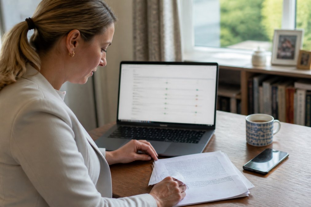 A person at a wooden desk in a bright home office comparing a laptop showing an online property case tracker with a printed document and a mobile phone beside a mug of tea