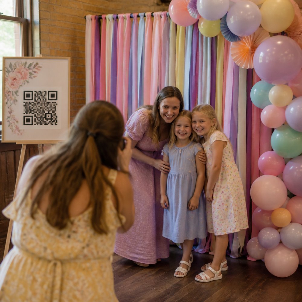 Colorful DIY photo booth backdrop with pastel streamers and balloons set up in bright party room, QR code poster displayed on easel to the side, guests smiling while posing in front of the setup