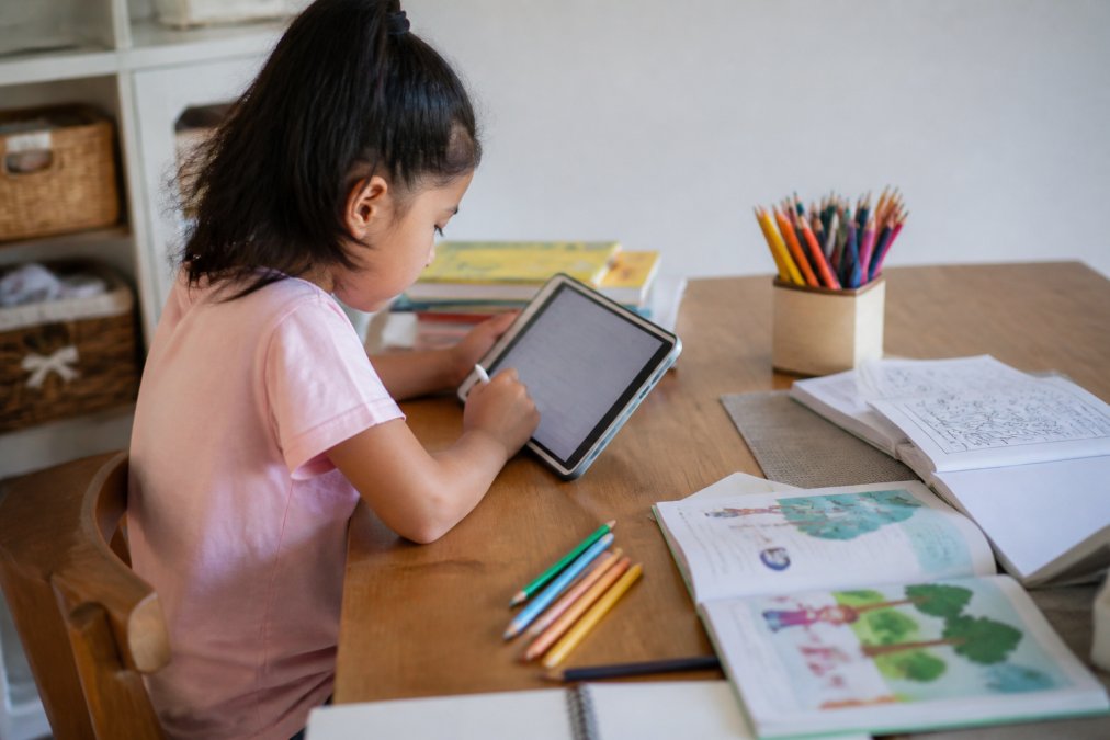 Child sitting at a desk with a tablet displaying a writing app, surrounded by paper, colored pencils, and activity books showing a blend of digital and hands-on creative work