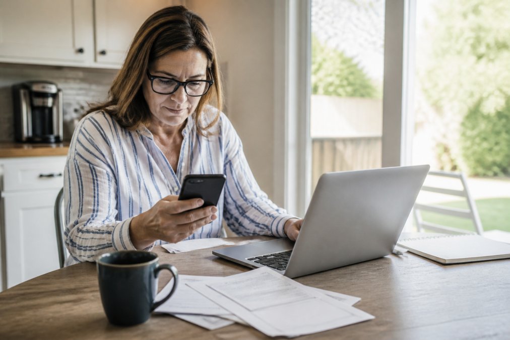 A person sitting at a kitchen table using a smartphone and laptop to review insurance policy documents, with a cup of coffee nearby and natural light coming through a window
