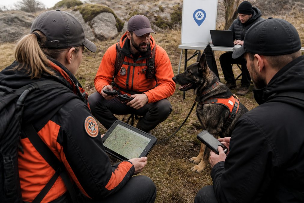 K9 handler and team members in the field using handheld GPS devices and tactical tablets during a live search and rescue operation, showing real-time coordination between handler, dog, and incident command center via digital mapping display