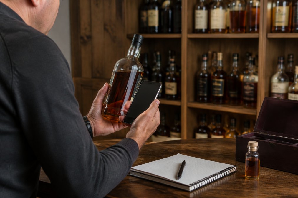 Collector's hands holding a bourbon bottle while using a smartphone to log details into a digital inventory app, with shelves of bottles visible in the background