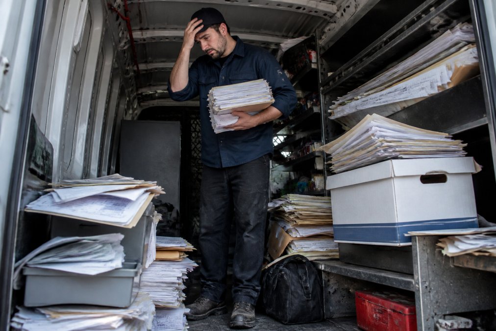 Field technician standing in a truck surrounded by stacks of paper work orders, folders, and filing boxes, looking overwhelmed