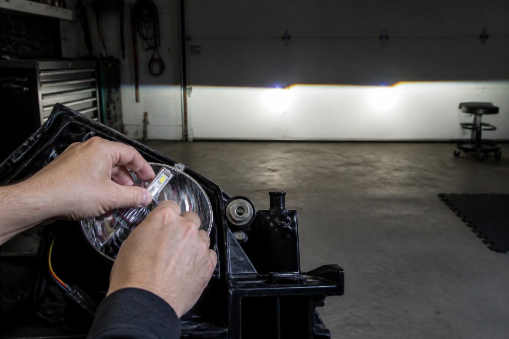 Close-up of a mechanic's hands adjusting an LED headlight bulb inside a vehicle's reflector housing, with a clear beam pattern visible on a garage wall in the background
