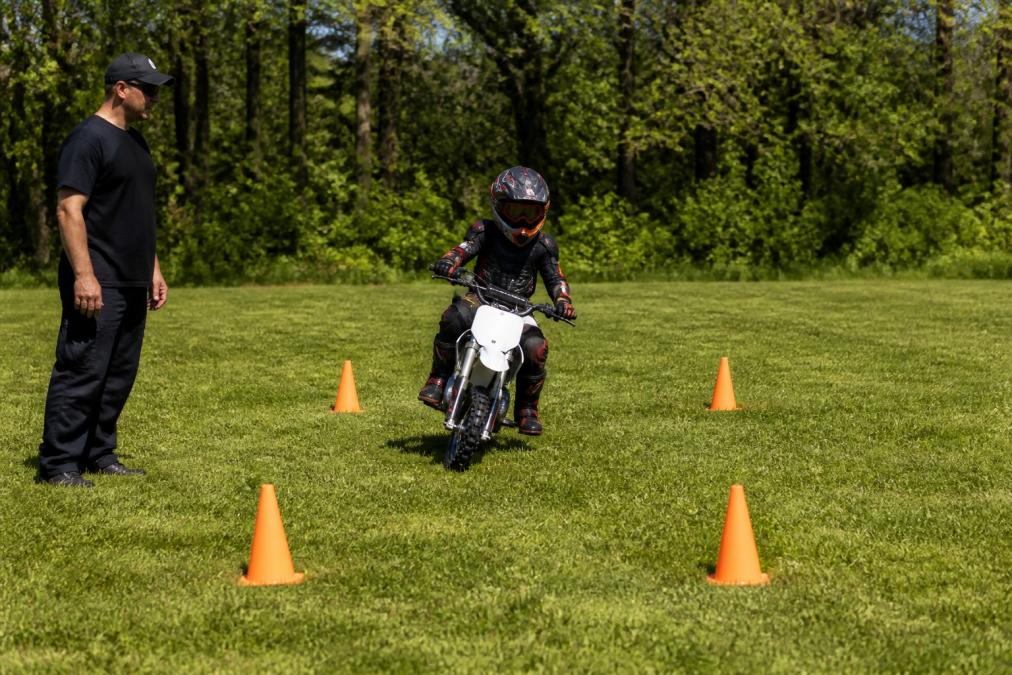Young rider practicing figure-eight riding drills on a dirt bike between orange cones on a grassy field