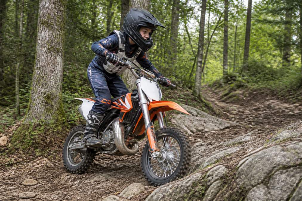 Young rider on a dirt bike demonstrating proper standing body position on a technical trail, showing elbows up, knees bent, weight centered over foot pegs, and head positioned forward looking through the turn