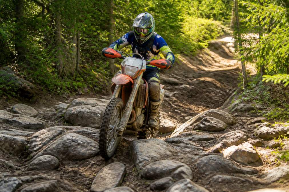 Dirt bike rider navigating rocky, rutted trail terrain with proper body positioning, weight distribution over the bike, and line selection through technical ground