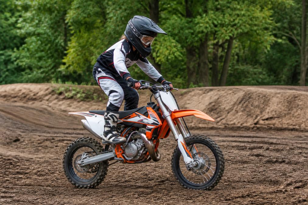 A young rider standing on the footpegs of a dirt bike, knees bent, elbows up, demonstrating proper body positioning on a dirt track with trees in the background