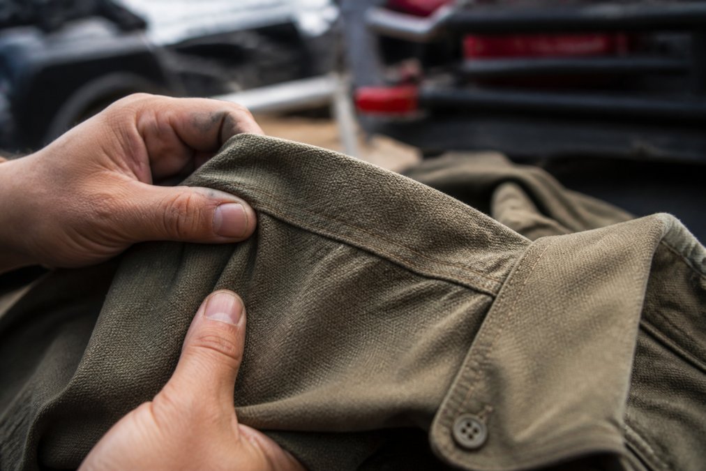 Close-up of hands examining heavyweight off-road apparel fabric, showing texture and reinforced stitching detail on a field shirt, natural lighting highlighting the weave pattern