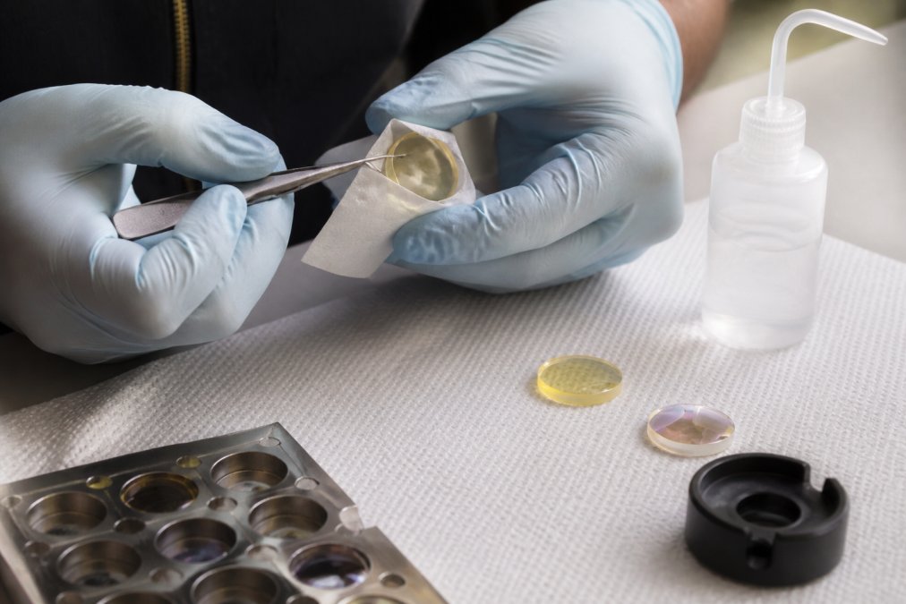 Professional technician's hands carefully cleaning a CO2 laser optics lens with optical-grade lens paper and cleaning solution, using precision tweezers on a clean workbench with proper lighting