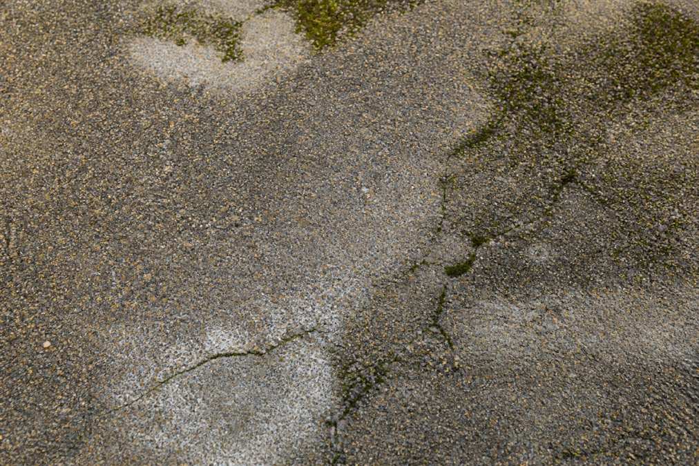 Close-up of weathered resin driveway surface showing visible cracks, moss growth patches, and discoloured areas from moisture damage and UV exposure in natural daylight