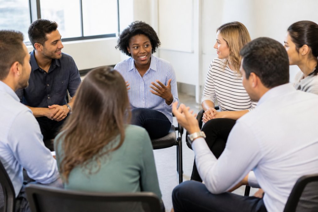 A diverse group of employees engaged in animated discussion during a role-playing exercise, sitting in a circle with engaged expressions and gestures, in a bright conference room with natural lighting