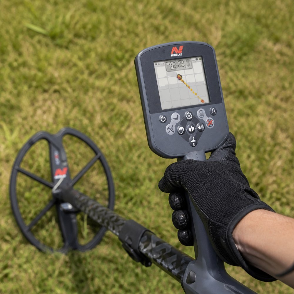 Close-up of a metal detector control unit display showing target ID numbers and discrimination settings, held by a detectorist's gloved hand outdoors on a sun-lit grassy field