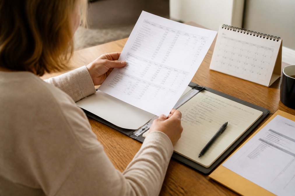 Person sitting at a desk reviewing printed lab reports and handwritten notes in a folder, with a pen and calendar visible, preparing documents for a healthcare consultation