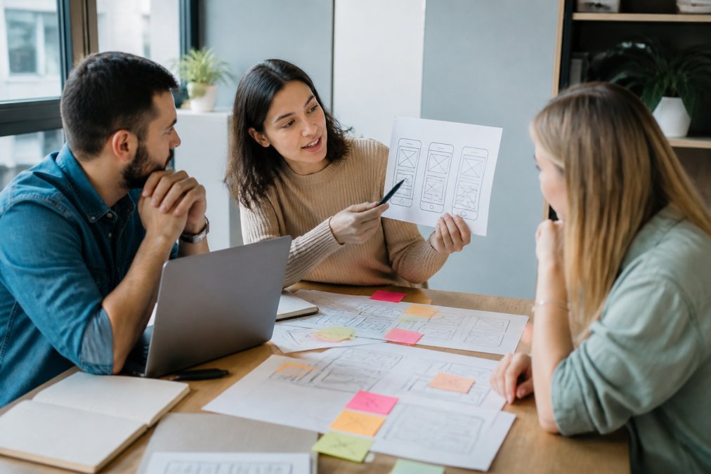 Diseñadora UX mostrando wireframes de diseño app a sus compañeros en una mesa de oficina con notas adhesivas