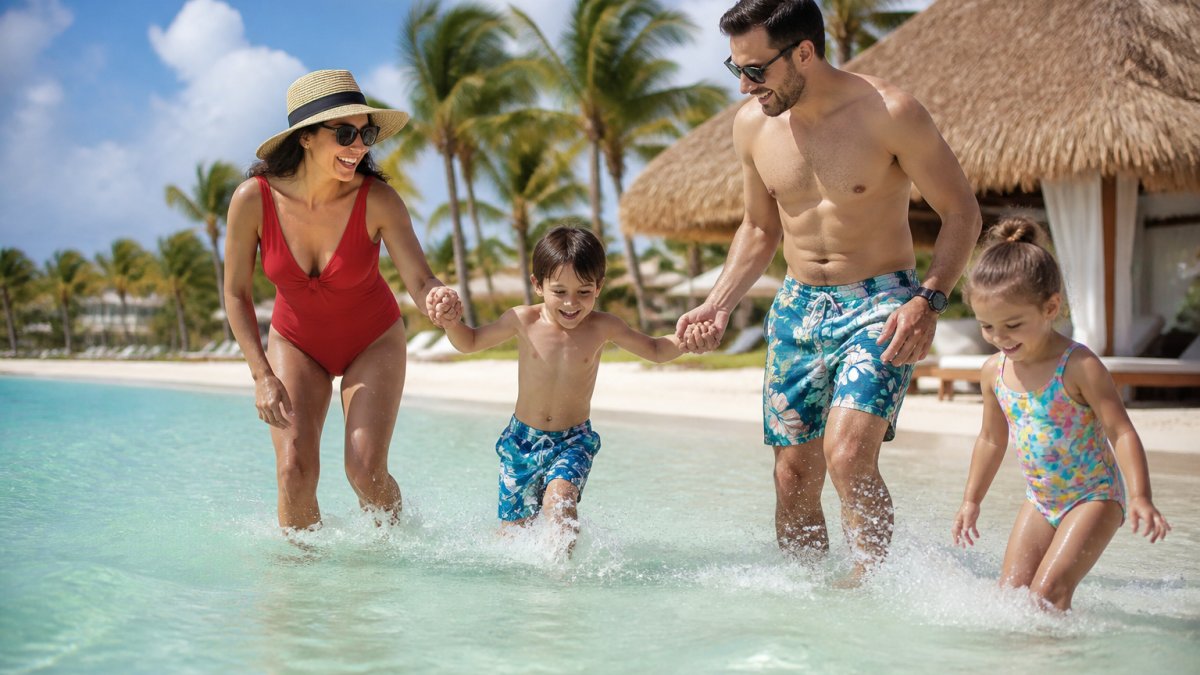 Family with young children playing in shallow turquoise water at a tropical beach resort, with palm trees and a thatched-roof cabana in the background