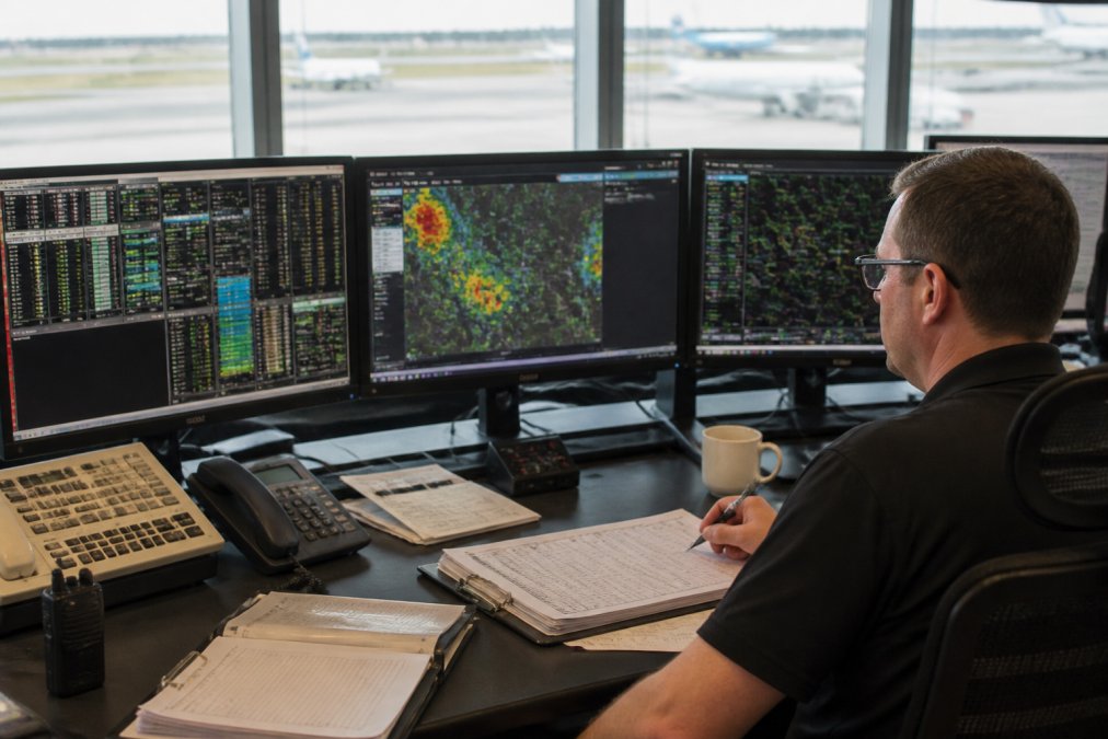 Aircraft dispatcher at a control room workstation with multiple monitors displaying flight data, weather maps, and communication systems, focused on coordinating flight operations