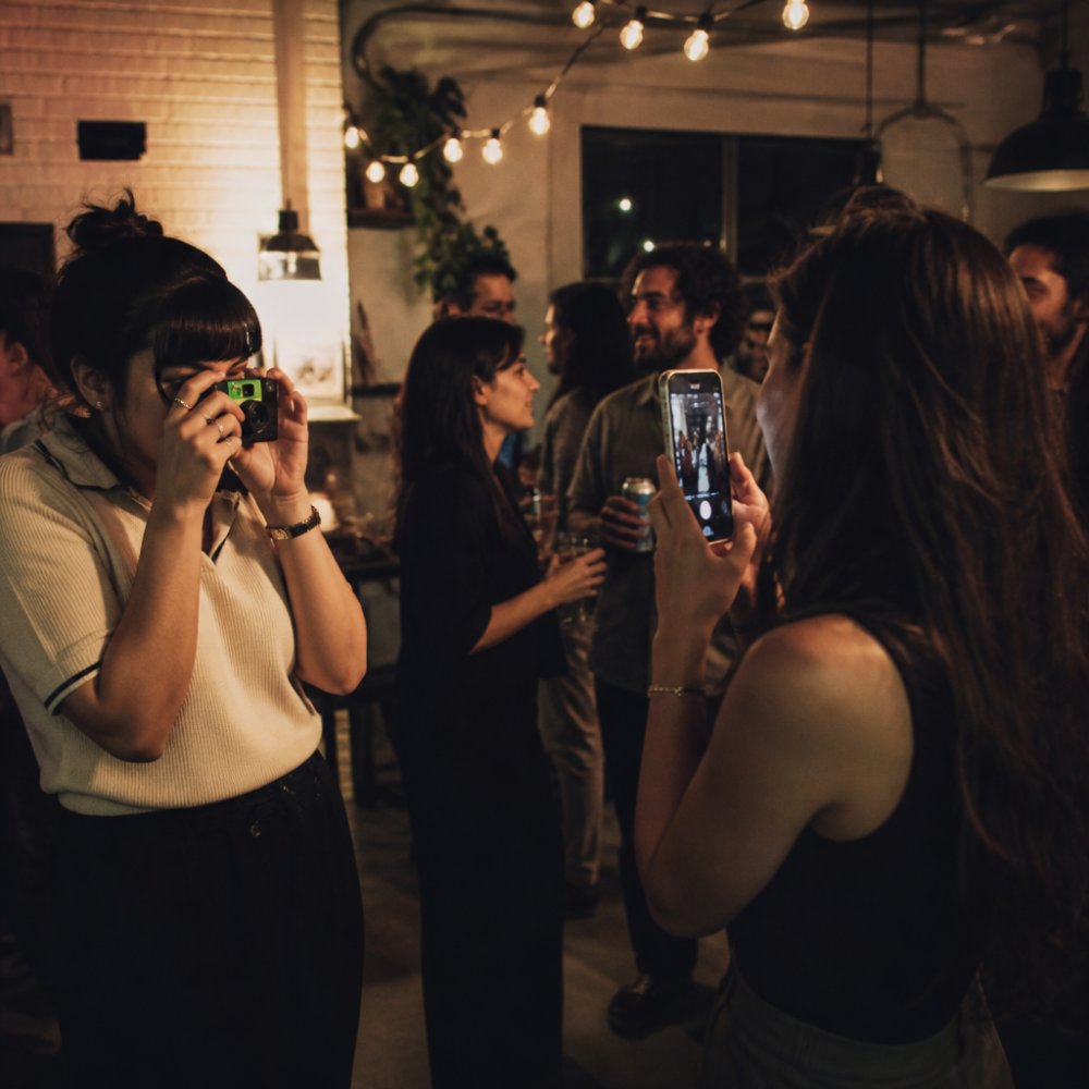 A lively indoor party scene with guests talking and laughing, one person holding a disposable film camera to their eye while another guest takes a photo with a smartphone, warm ambient lighting, candid atmosphere