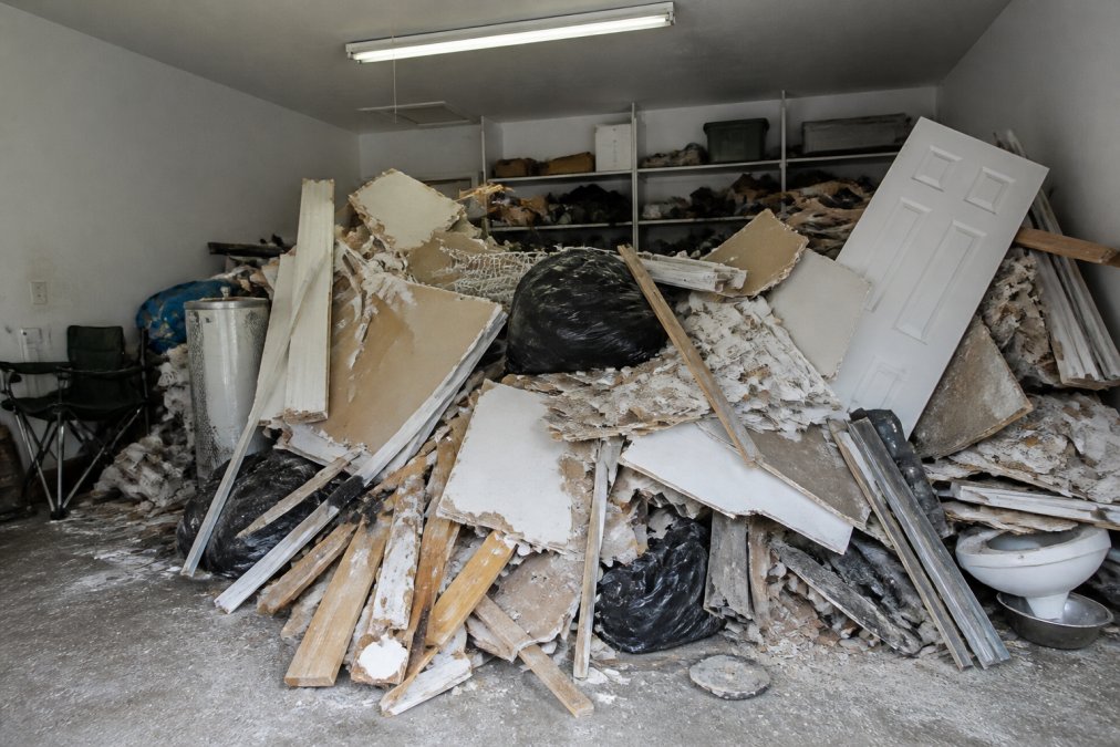 Garage filled with renovation debris piled up showing drywall pieces, wood scraps, metal fixtures, concrete chunks, and mixed materials stacked against walls under fluorescent lighting