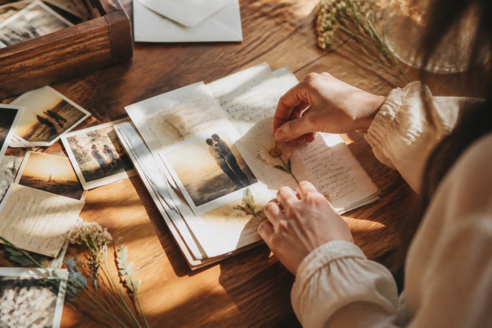 Hands arranging handwritten letters, vintage photographs, and pressed flowers on a wooden table with warm natural sunlight streaming across the surface, showing the assembly of a sentimental memory collection