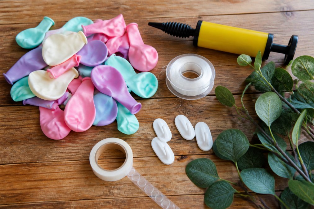 Flat lay of balloon garland supplies arranged on wooden table: assorted latex balloons in pastel colors, yellow hand pump, clear monofilament line spool, white command hooks, double-sided balloon tape roll, and faux eucalyptus sprigs