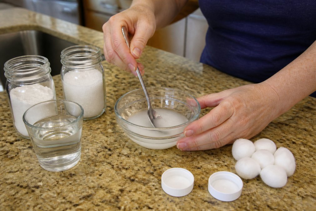 Close-up of hands mixing borax, sugar, and warm water in a small bowl on a kitchen counter, with cotton balls and bottle caps nearby