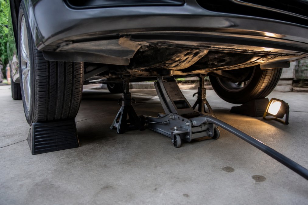 Close-up of a floor jack and jack stands properly supporting a vehicle on level concrete driveway, with safety chocks wedged against the front tire and work light illuminating the undercarriage