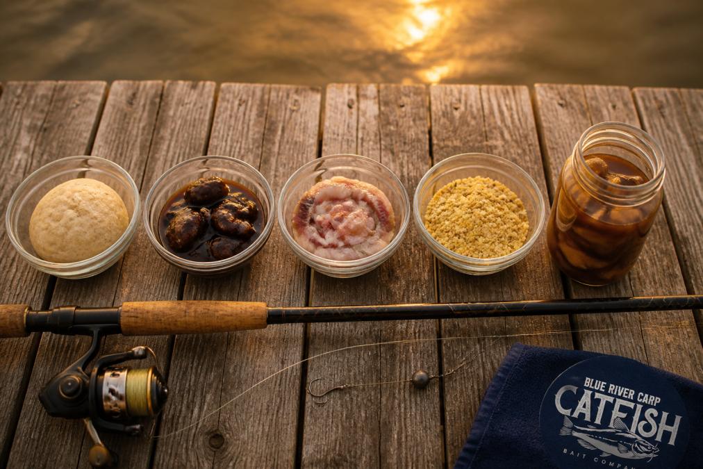 Overhead shot of a wooden dock at golden hour with five small bowls of homemade catfish bait lined up: a dough ball, a molasses-coated chunk, a cut bait strip, a cornmeal mix, and a jar of marinated bait, with a fishing rod resting nearby