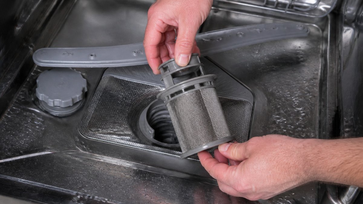 Close-up of hands removing a dishwasher filter from the bottom of the appliance, showing the exact location and removal technique with water droplets visible