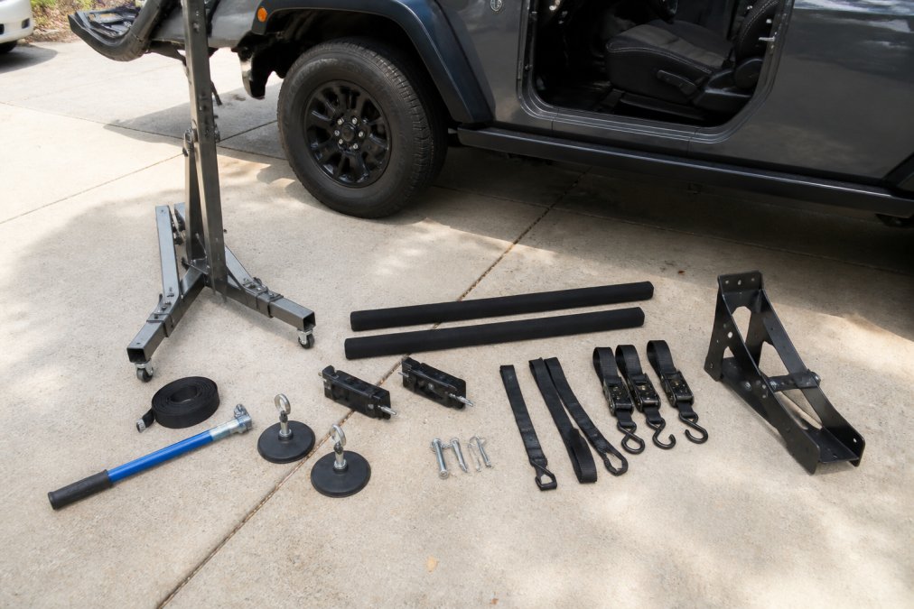 A Jeep Wrangler owner standing beside their vehicle in a sunlit driveway, using a standalone hardtop removal tool positioned over the hood to lift the hardtop clear of the body