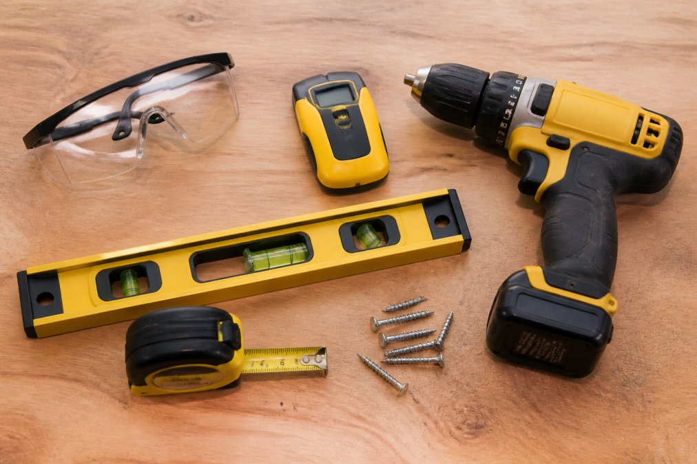 Overhead flat-lay arrangement of essential DIY cabinet installation tools including cordless drill, laser level, stud finder, tape measure, square, clamp, and safety glasses arranged on a wooden workbench
