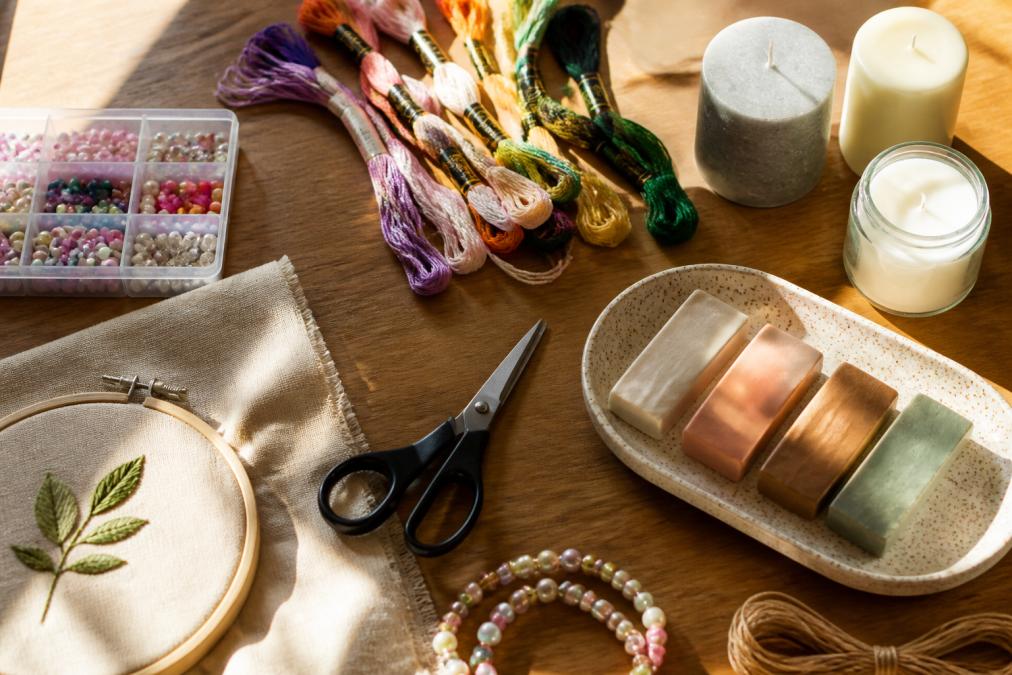 Flat lay of craft supplies including colorful embroidery floss, beads, candles, clay, and scissors arranged on a wooden table with natural sunlight streaming across