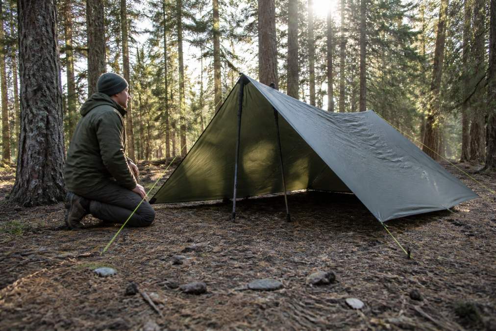 A hiker in rugged woodland kneeling beside a completed A-frame poncho tarp shelter, two trekking poles supporting the ridgeline, paracord guy lines tensioned to stakes, morning light filtering through trees