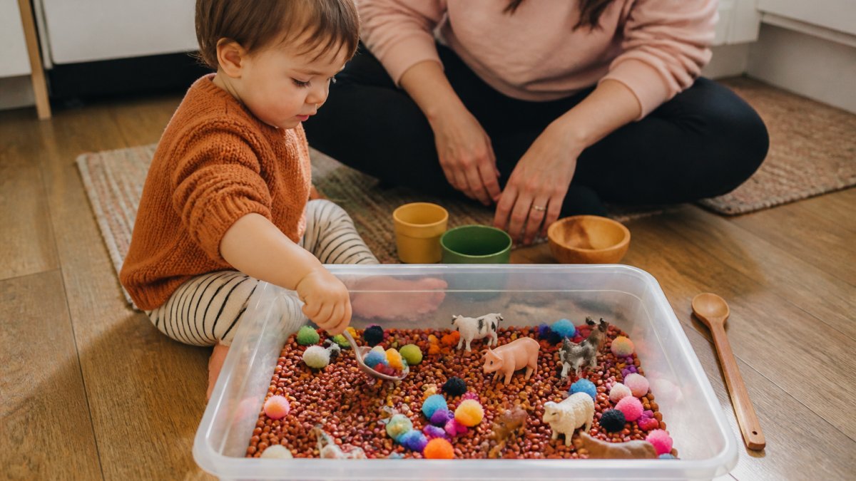 A toddler sitting on a kitchen floor, scooping colourful pom-poms and plastic animals from a shallow plastic tub filled with dried beans, wooden spoon and small cups nearby, natural light, warm home setting, parent's hands visible in the background