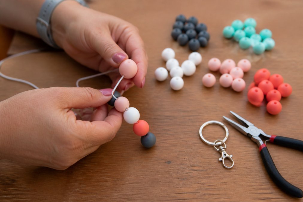 Close-up of hands threading 15 mm silicone beads onto a stretchy cord on a wooden craft table, with small piles of colorful beads, a lobster clasp keyring, and pliers nearby