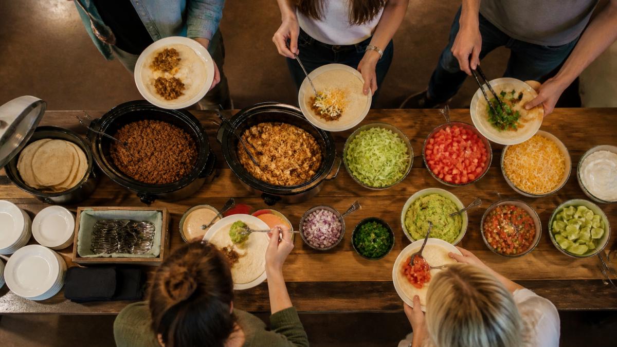 Overhead view of a fully arranged DIY taco bar with warm tortillas in a tortilla warmer, seasoned ground beef and shredded chicken in slow cookers, colorful bowls of shredded lettuce, diced tomatoes, shredded cheese, sour cream, guacamole, and fresh salsa arranged in rows, with serving utensils and small plates stacked at the start of the line, and guests serving themselves