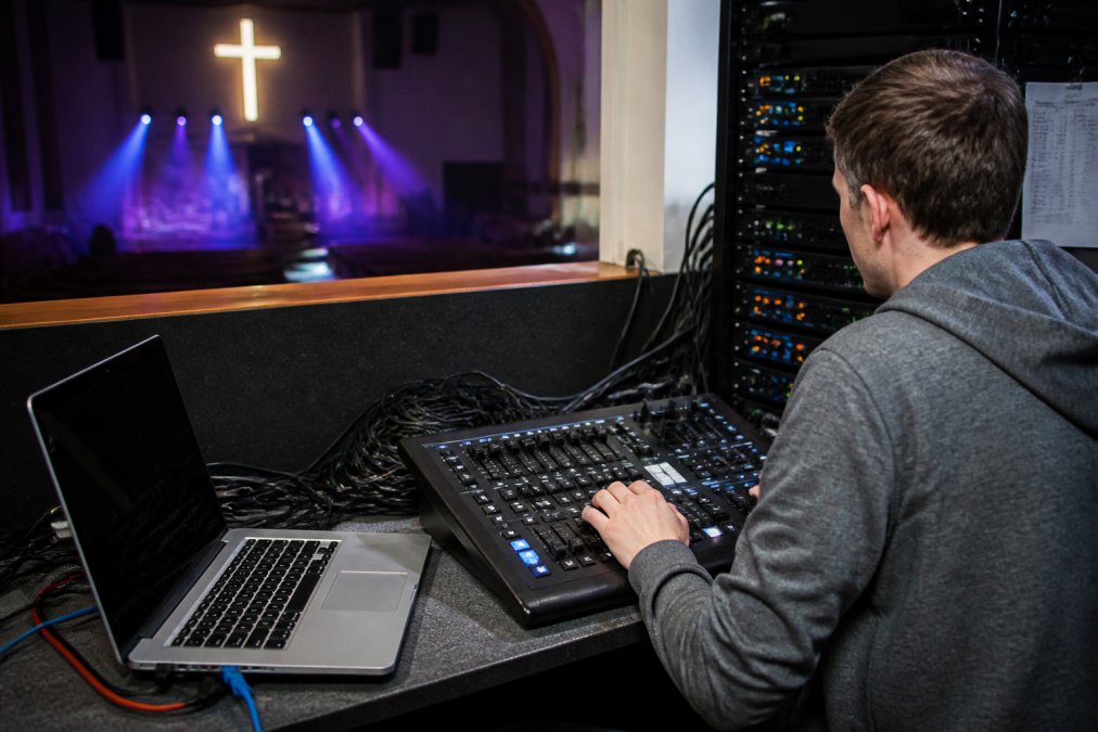 A volunteer at a church tech booth adjusting a lighting console with a laptop showing DMX software, cables and a dimmer rack visible in a compact booth space