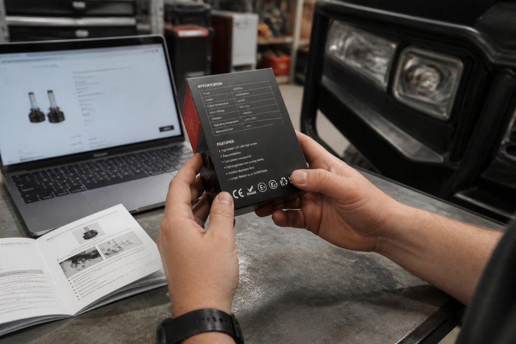 Close-up of a person inspecting the back of an LED headlight bulb box for compliance markings, with a vehicle owner's manual and a laptop showing a product page on a garage workbench