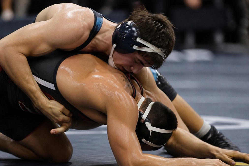Two wrestlers in close contact during a match on a padded mat, showing direct skin-to-skin grappling with sweat visible