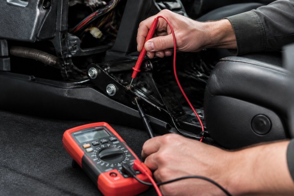 Automotive technician using a digital multimeter to test grounding continuity on a vehicle's interior component, with probe tips making contact on metal surfaces and the meter displaying resistance readings