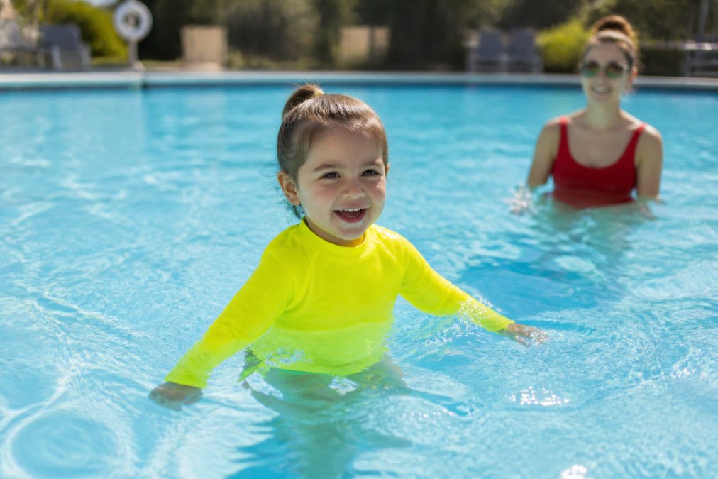 Young child wearing neon high-visibility swimsuit in bright pool setting, easily visible against the blue water, with a parent or lifeguard visible in the background supervising closely