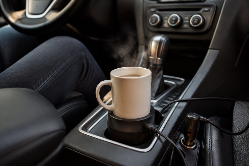 Portable electric mug warmer plugged into a car 12V adapter with steam rising from a ceramic mug inside a cup holder