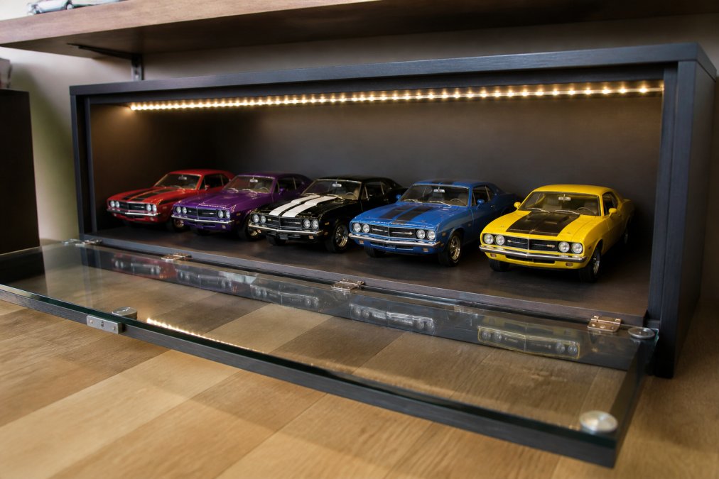 A collector's shelf holding several different 1:18 scale muscle car models from various manufacturers, arranged inside a well-lit display diorama with a sliding front panel pulled open