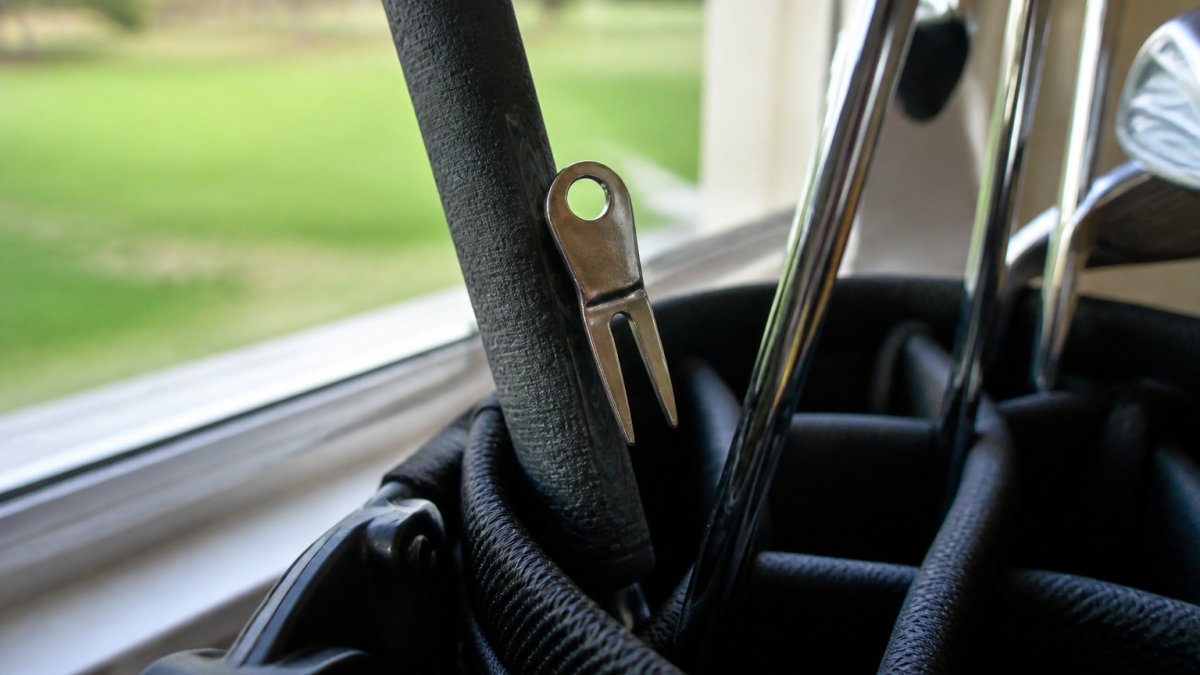 Close-up of an open golf bag with a putter standing upright and a metal divot repair tool resting against the putter grip, prongs touching the rubber, soft daylight from a clubhouse window