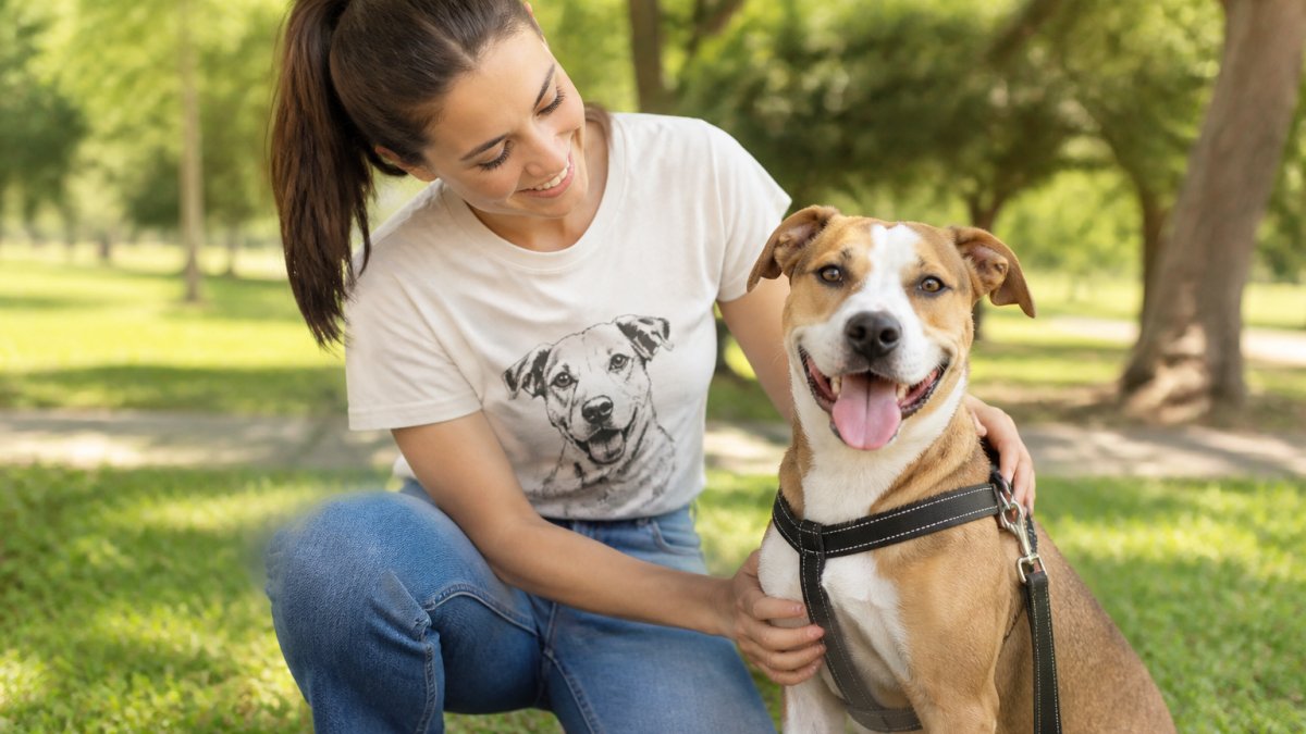 A person wearing a dog-themed t-shirt kneeling and smiling with a rescue dog in a sunny park