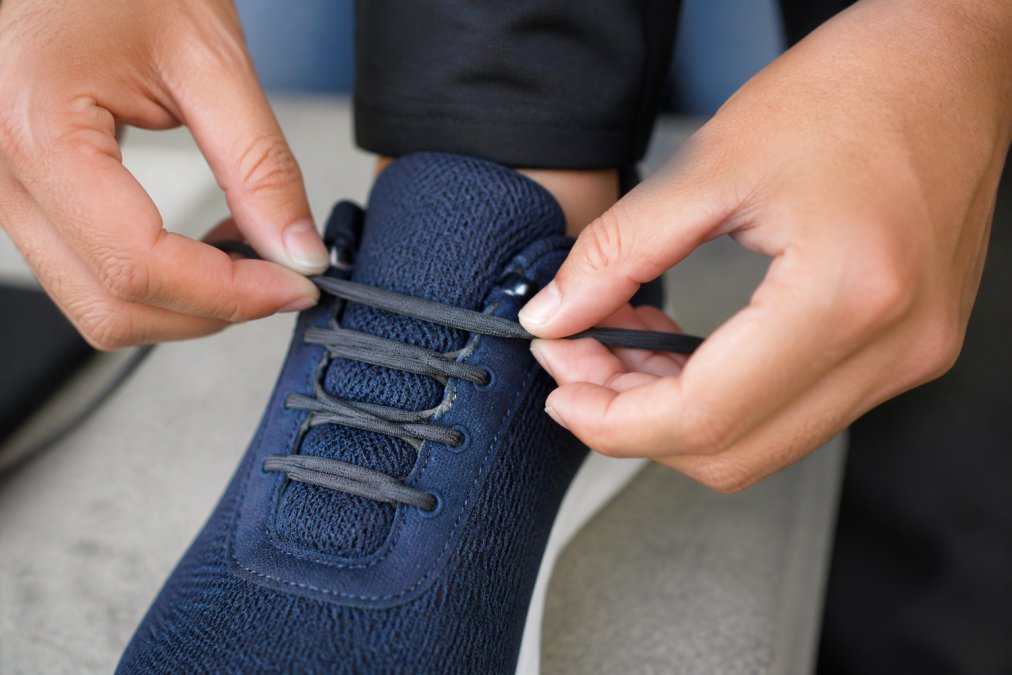 Close-up of hands demonstrating elastic lace installation through shoe eyelets, showing the stretchy material being threaded through on a running sneaker with natural lighting